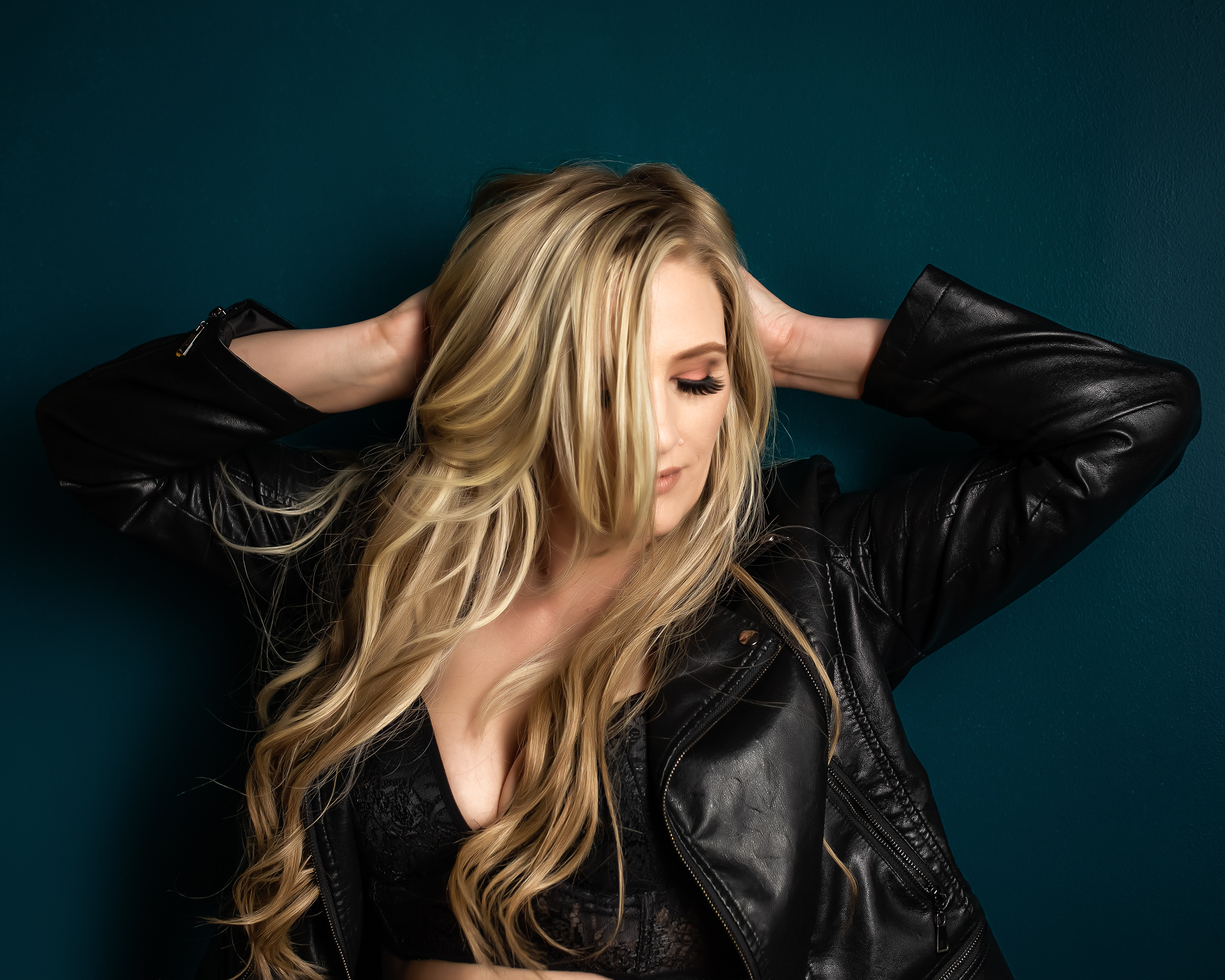 Woman wearing a black leather jacket poses against a teal wall with her arms behind her head and her hair covering her face.